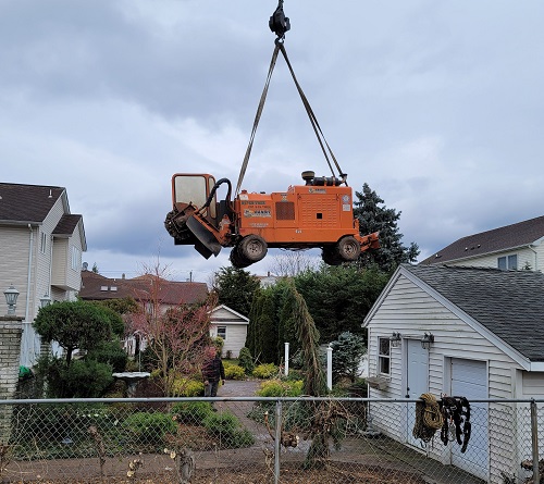 Aspen Tree Service Inc.'s crane lifting a stump grinder in the air over a chainlink fence to reach a confined back yard in Clifton, NJ.