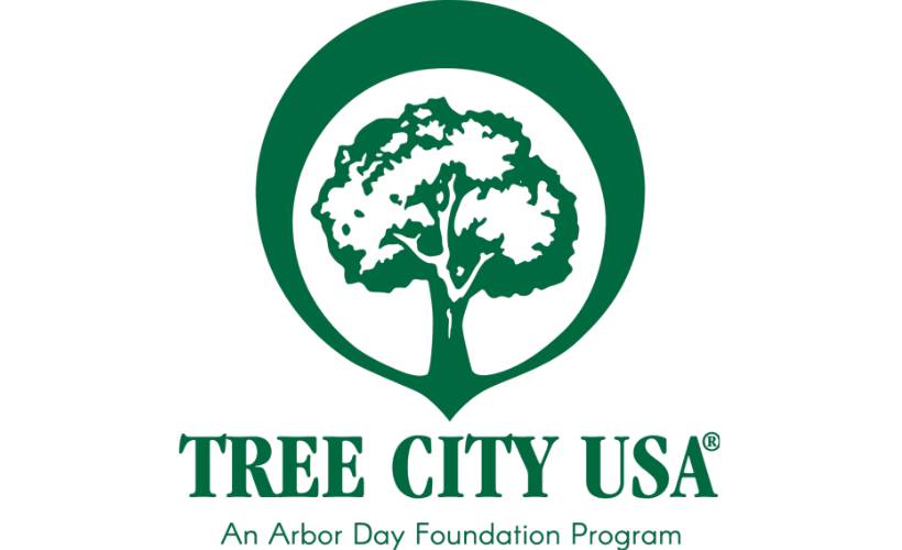 A green Tree City USA logo with a circle surrounding a drawing of a green tree and text reading An Arbor Day Program beneath.