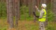 Arborist in safety vest and hard hat inspecting tree trunk in a wooded area while holding a laptop.